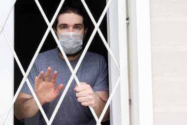 Tek kullanımlık maskeli, siyah saçlı, sakallı ve gri tişörtlü bir erkek manken. Coronavirus hastası evde karantina ve hastalık önleme altında kalıyor. Bir insanın gözlerinde olumsuz ve olumsuz duygular
