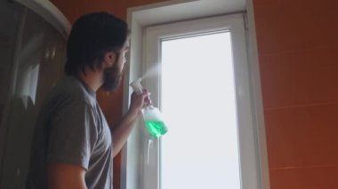 Gri tişörtlü ve sakallı bir adam evin pencerelerini temizlemek için temizlik maddesi ve sünger kullanıyor.