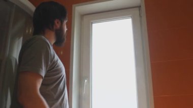 Gri tişörtlü ve sakallı bir adam evin pencerelerini temizlemek için temizlik maddesi ve sünger kullanıyor.