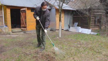 Koronavirüsten korunmak için tek kullanımlık maske takan genç adam, ağaç yapraklarından bahar temizliği yapıyor. Rüzgar saçlarını değiştiriyor. Doğa koruma kavramı ve çöpten orman temizliği kavramı.