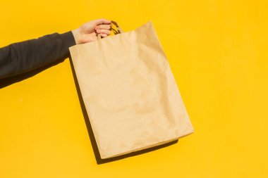 Sarı arka planda paket almak için kahverengi boş el işi kağıt torbası ile erkek elini kapat. Paketleme şablonu modeli. Coronavirus sırasında yemek teslimatı, konsept. Reklamcılık için alanı kopyala