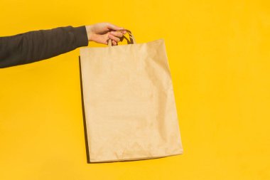 Sarı arka planda paket almak için kahverengi boş el işi kağıt torbası ile erkek elini kapat. Paketleme şablonu modeli. Coronavirus sırasında yemek teslimatı, konsept. Reklamcılık için alanı kopyala