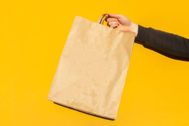 Sarı arka planda paket almak için kahverengi boş el işi kağıt torbası ile erkek elini kapat. Paketleme şablonu modeli. Coronavirus sırasında yemek teslimatı, konsept. Reklamcılık için alanı kopyala