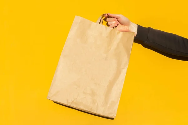 Sarı arka planda paket almak için kahverengi boş el işi kağıt torbası ile erkek elini kapat. Paketleme şablonu modeli. Coronavirus sırasında yemek teslimatı, konsept. Reklamcılık için alanı kopyala