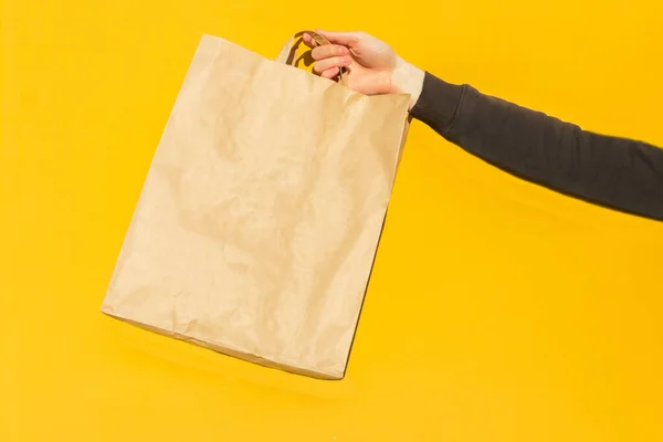 黄色の背景に持ち帰り用の手茶色の空の空白のクラフト紙袋に男性のホールドを閉じます パッケージテンプレートのモックアップ コロナウイルスにおける食品配送サービスの概念です 広告のコピースペース — ストック写真