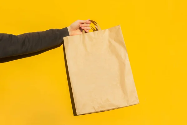 Sarı arka planda paket almak için kahverengi boş el işi kağıt torbası ile erkek elini kapat. Paketleme şablonu modeli. Coronavirus sırasında yemek teslimatı, konsept. Reklamcılık için alanı kopyala