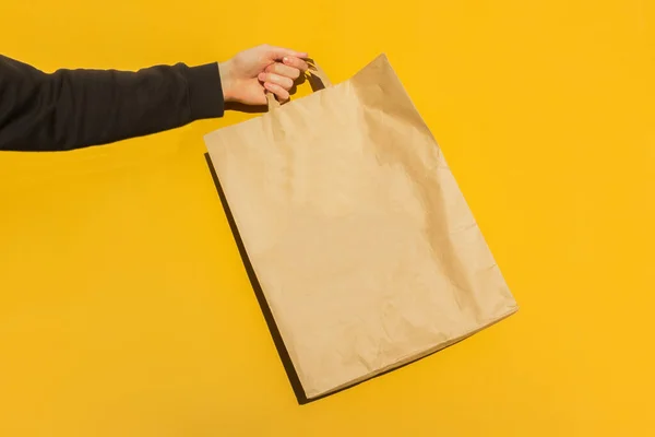 黄色の背景に持ち帰り用の手茶色の空の空白のクラフト紙袋に男性のホールドを閉じます パッケージテンプレートのモックアップ コロナウイルスにおける食品配送サービスの概念です 広告のコピースペース — ストック写真