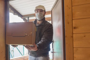 Koruyucu maskeli kurye karantina, salgın hastalık salgını ve koronavirüs salgını altında paket teslim eder. Beyaz şapkalı bir erkek kuryenin tuttuğu karton kutunun yakın çekimi.