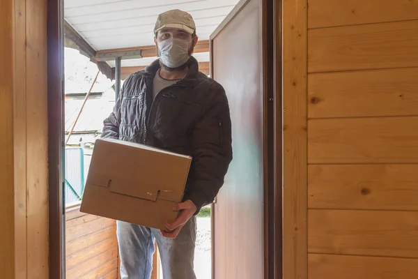 Koruyucu maskeli kurye karantina, salgın hastalık salgını ve koronavirüs salgını altında paket teslim eder. Beyaz şapkalı bir erkek kuryenin tuttuğu karton kutunun yakın çekimi.