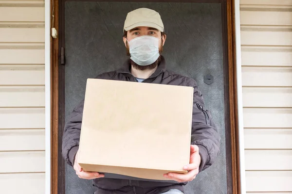 Koruyucu maskeli kurye karantina, salgın hastalık salgını ve koronavirüs salgını altında paket teslim eder. Beyaz şapkalı bir erkek kuryenin tuttuğu karton kutunun yakın çekimi.