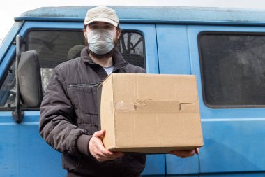 Koruyucu maskeli kurye, karantina altında araba teslimatı, hastalık salgını, koronavirüs salgını durumları. Beyaz şapkalı bir erkek kuryenin tuttuğu karton kutunun yakın çekimi.