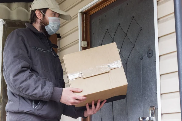 Courier in protective mask delivers parcel, Home service delivery under quarantine, coronavirus pandemic conditions. close-up of cardboard box holding by a male courier in a white cap