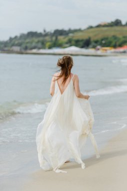 Süt renkli uzun elbiseli güzel bir kız sahilde yürüyor ve denizin arka planında rıhtımda yürüyor..