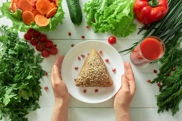 Coarse bun on the background of vegetables. Female hands reach for a plate showing the correct selection of products for weight loss.