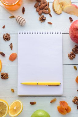 Elmalar, kiviler, kurutulmuş meyveler, portakallar ve elmalarla çevrili kalemli defter. Sağlıklı beslenme ve alışveriş listesi kavramı.