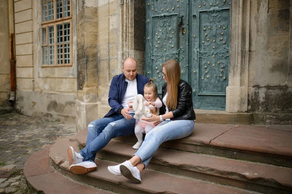 Parents and their daughter are sitting on the steps of an old church.