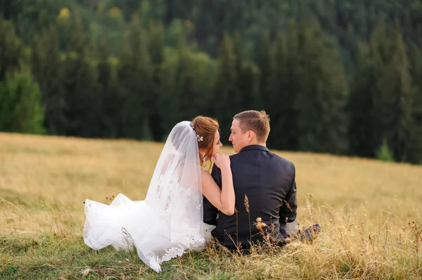 在山上拍婚纱照新婚夫妇背靠在草地上坐着互相望着 — 图库照片