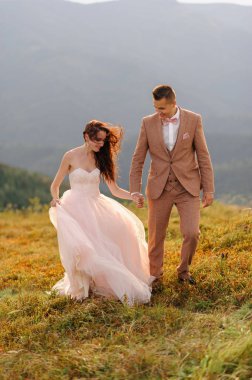 Gelin ve damat yan yana gidiyor. Gün batımı. Sonbahar dağlarının arka planında düğün fotoğrafı. Güçlü bir rüzgar saç ve elbise şişirir..