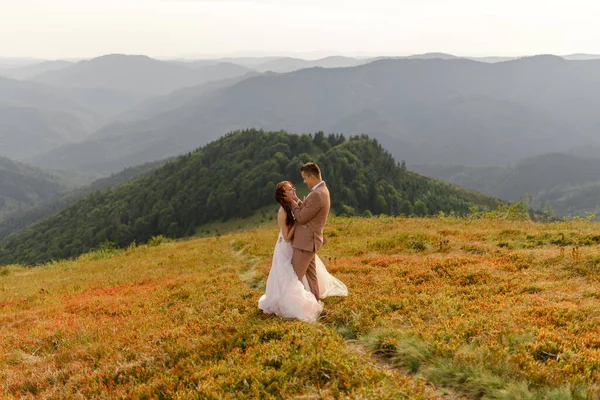 新娘和新郎拥抱在一起 日落了秋山背景下的婚纱照 大风使头发和衣服膨胀起来 后续行动 — 图库照片