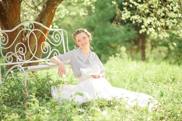 若い美しい女性は リンゴの木の開花を背景にベンチの近くに地面に座っています ベンチに本があります 白い鳩を手にしてる — ストック写真