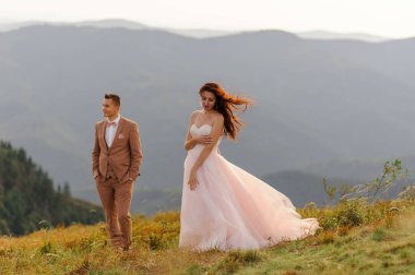 Gelin ve damat birbirlerine nazikçe sarılıyorlar. Gün batımı. Sonbahar dağlarının arka planında düğün fotoğrafı. Güçlü bir rüzgar saç ve elbise şişirir.. 