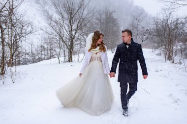 Gelin ve damat bir kış ormanının arka planında el ele yürüyorlar. Kar yağıyor.