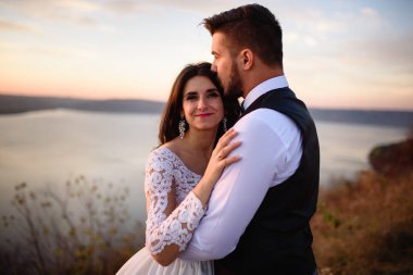 Gelin ve damat gün batımında gölün arka planında sarılıyorlar..