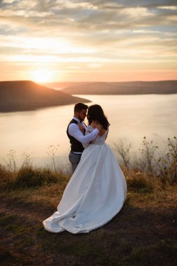 Gelin ve damat gün batımında gölün arka planında sarılıyorlar..