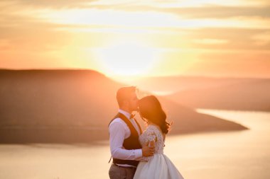 Gelin ve damat gün batımında gölün arka planında sarılıyorlar..