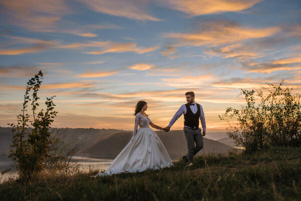 The groom leads the bride by the hand during sunset.