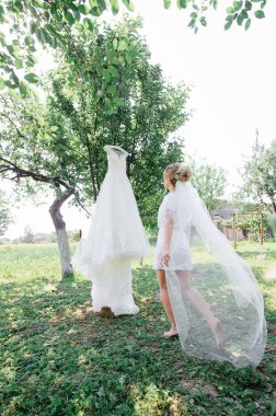 Gelinliğinin yanında poz veren güzel gelin peignoir. Elbise bahçedeki bir ağaca asılı..