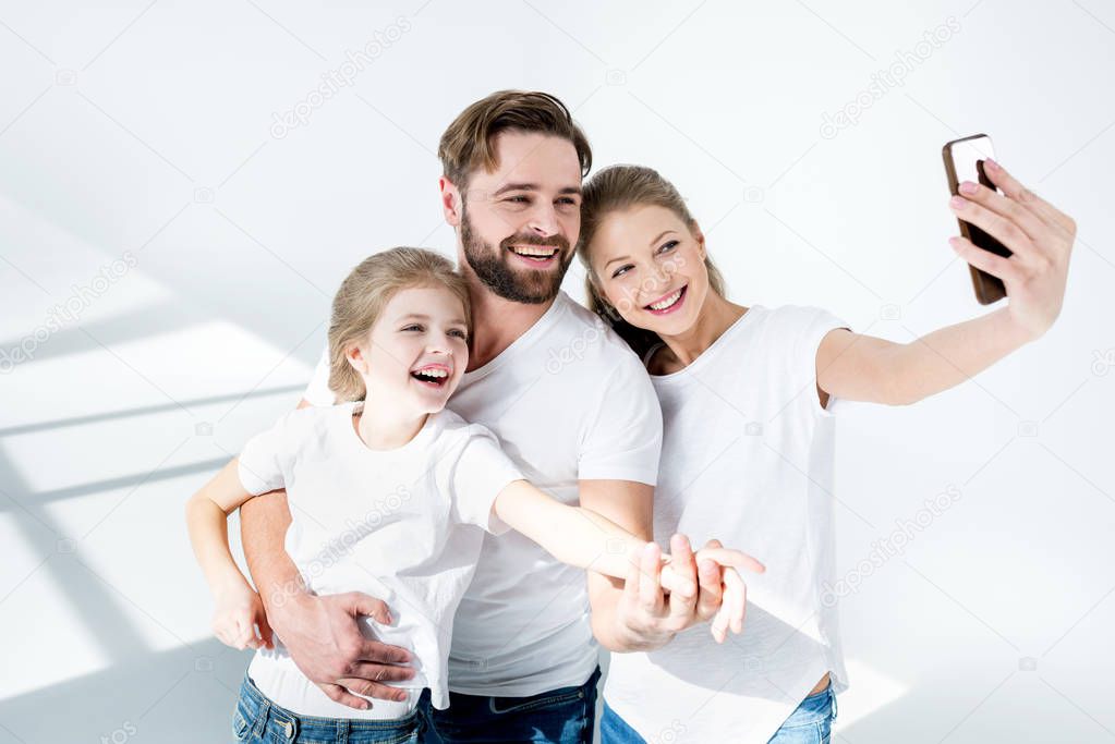 Happy young family in white t-shirts taking selfie with smartphone in studio