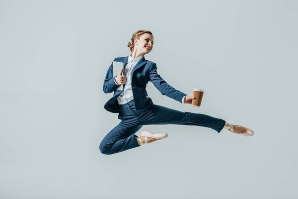 businesswoman in suit and ballet shoes jumping with coffee and digital tablet, isolated on grey