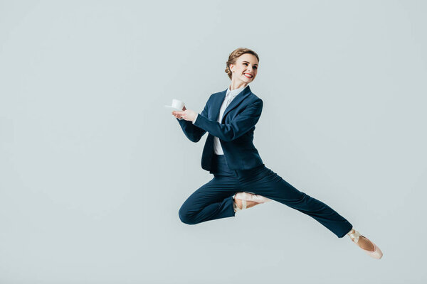 деловая женщина в костюме и балетной обуви, прыгающая с чашкой кофе, изолированная на сером
