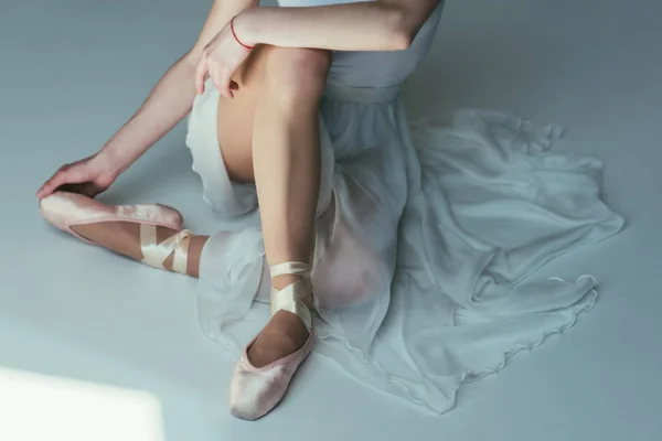 Обрезанный вид нежной балерины, сидящей в белом платье и балетной обуви — стоковое фото