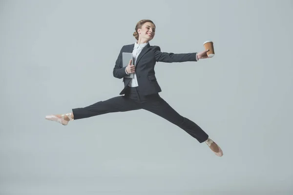 Femme d'affaires en costume et chaussures de ballet sautant avec café et tablette, isolé sur gris — Photo de stock