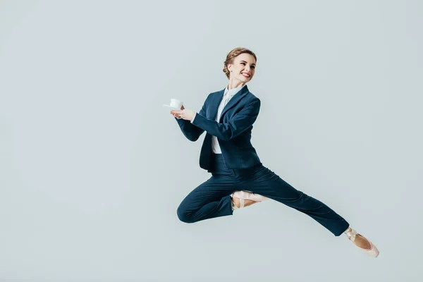 Donna d'affari in giacca e cravatta e scarpe da ballo che salta con una tazza di caffè, isolata su grigio — Foto stock