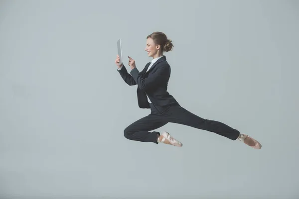 Geschäftsfrau in Anzug und Ballettschuhen springt und nutzt digitales Tablet, isoliert auf grau — Stockfoto