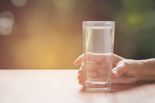 Mano sosteniendo un vaso de agua pura. Mano sosteniendo un vaso de agua en la mesa con fondo natural . — Foto de Stock