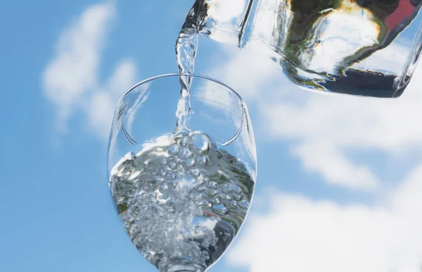 Θηλυκό χέρι ρίχνει νερό από το μπουκάλι στο γυαλί με μπλε ουρανό φόντο. — Φωτογραφία Αρχείου