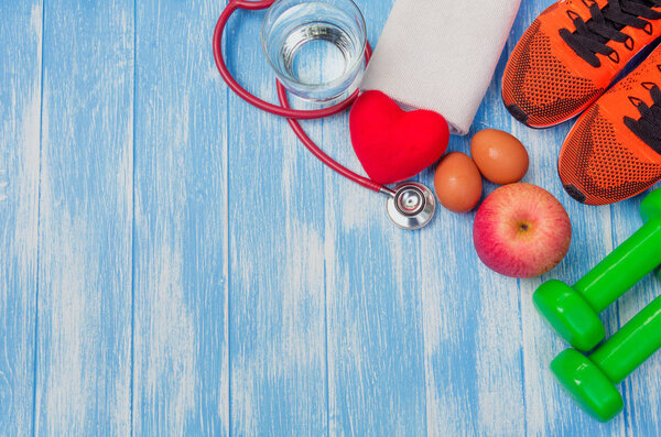 Top view of sportswear, sports equipment, dumbbells and stethoscope on blue and wooden table.