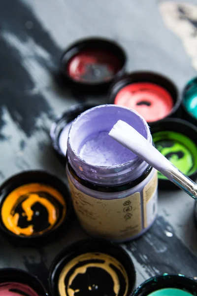 Jar with a violet paint and a brush, below a multi-colored jars