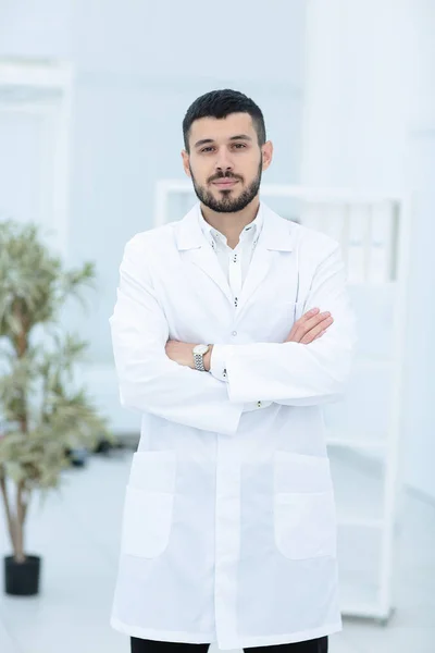S úsměvem lékař pózuje s rukama zkříženýma v kanceláři, má na sobě zdravotnický personál na pozadí — Stock fotografie