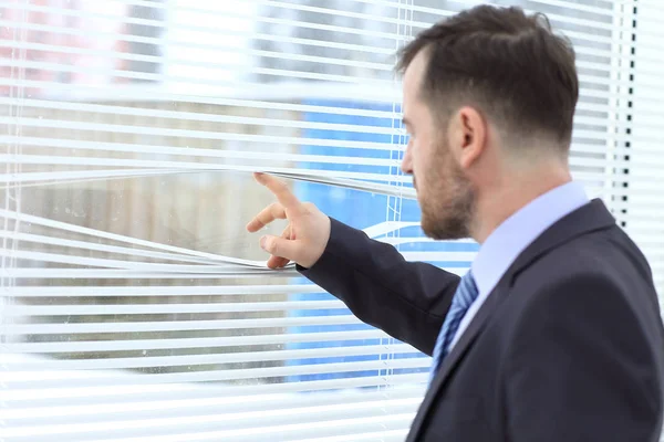 Young and handsome businessman looking out of the window through the blinds — Stock Photo, Image