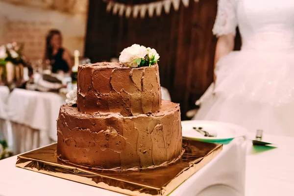 O bolo de casamento de chocolate de dois níveis é decorado com flores brancas e fica no suporte dourado, na mesa branca. . — Fotografia de Stock