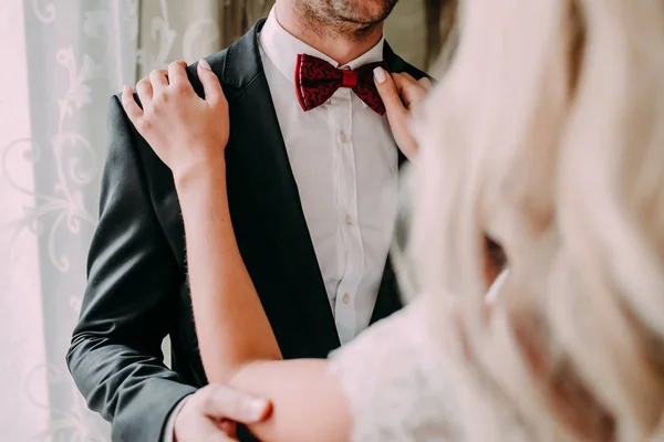 Charmante mariée fixe noeud papillon rouge sur le cou des mariés pendant qu'ils se tiennent près de la fenêtre. Mariage . — Photo
