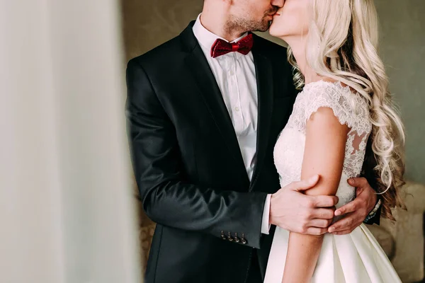 Los recién casados felices se besan en interiores. Una novia y un novio se paran cerca de la ventana. Boda . — Foto de Stock