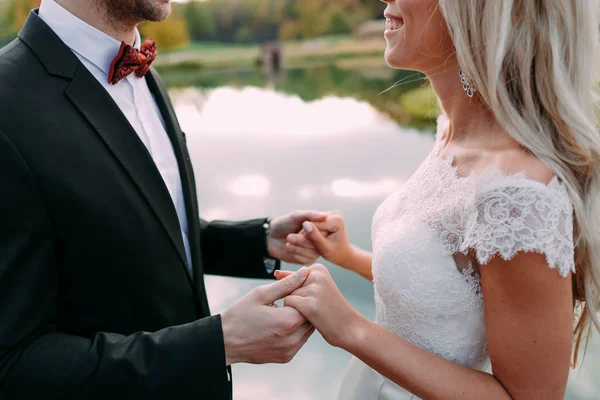 Boda. La novia y el novio están tomados de la mano y sonriendo. Vista lateral de la joven pareja de pie cara a cara y mirándose mientras están de pie contra el lago . — Foto de Stock
