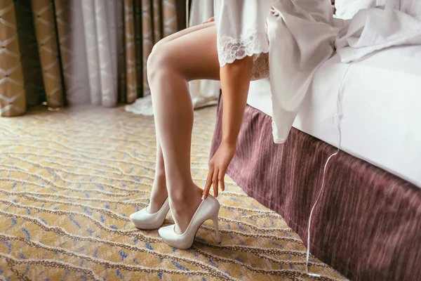 Bride in a silk robe with beautiful slim legs is putting on bridal shoes. Wedding morning preparation