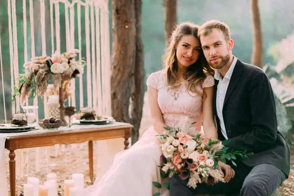 Los novatos felices miran a un fotógrafo. Hombre y mujer vestida de fiesta se sientan en las piedras cerca de la decoración de la boda en estilo boho. Una ceremonia al aire libre. Cierre — Foto de Stock